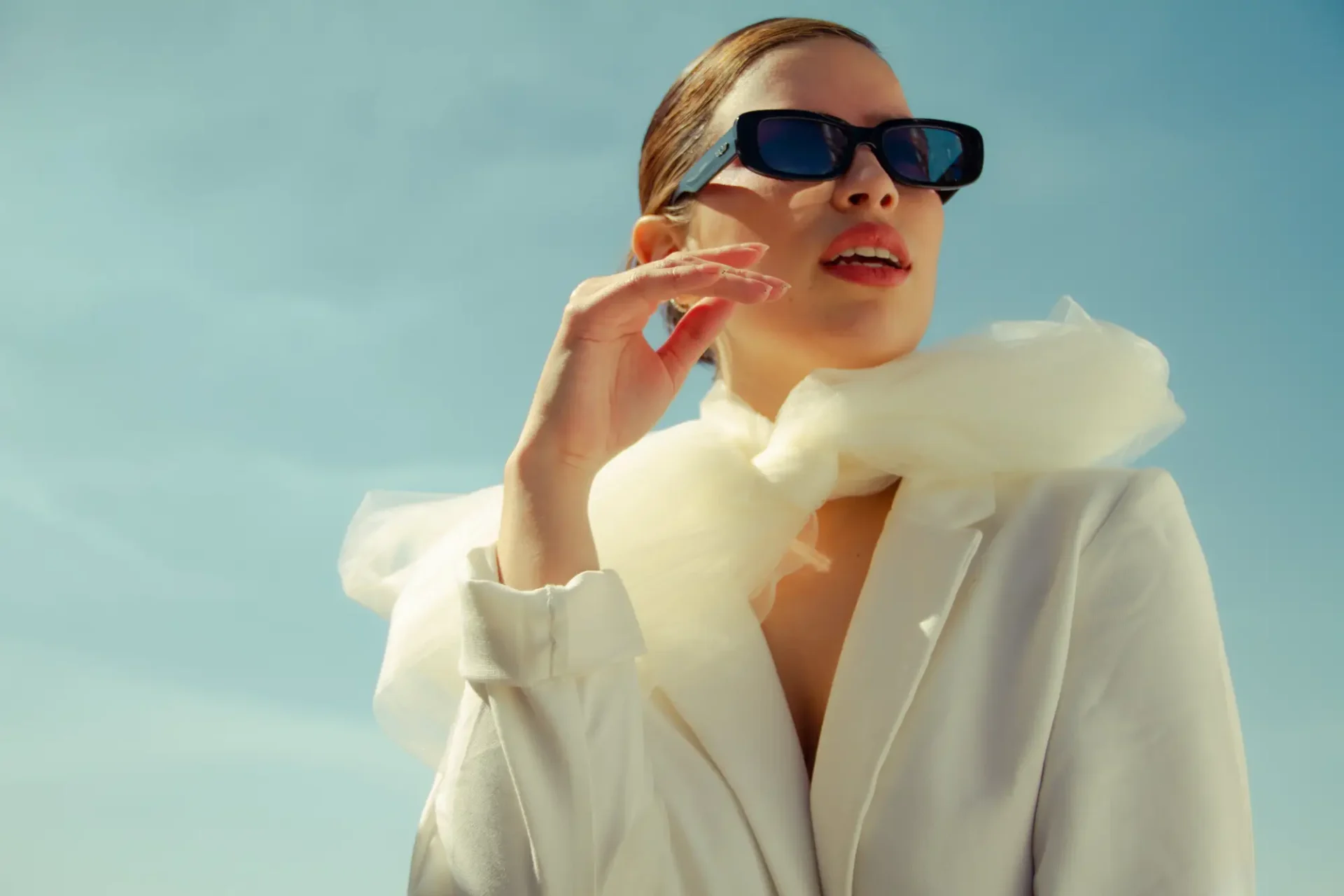 A woman in sunglasses and white coat holding her hand to the side of her face.